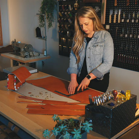 Woman measuring and cutting leather pieces on a workbench with leathercraft tools and supplies organized nearby.