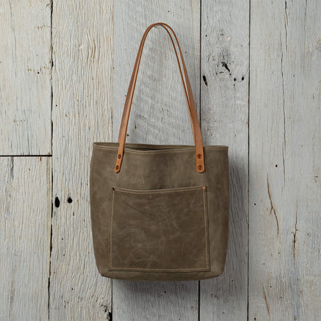 Handmade leather tote bag with front pocket and natural leather straps against a rustic wooden background.