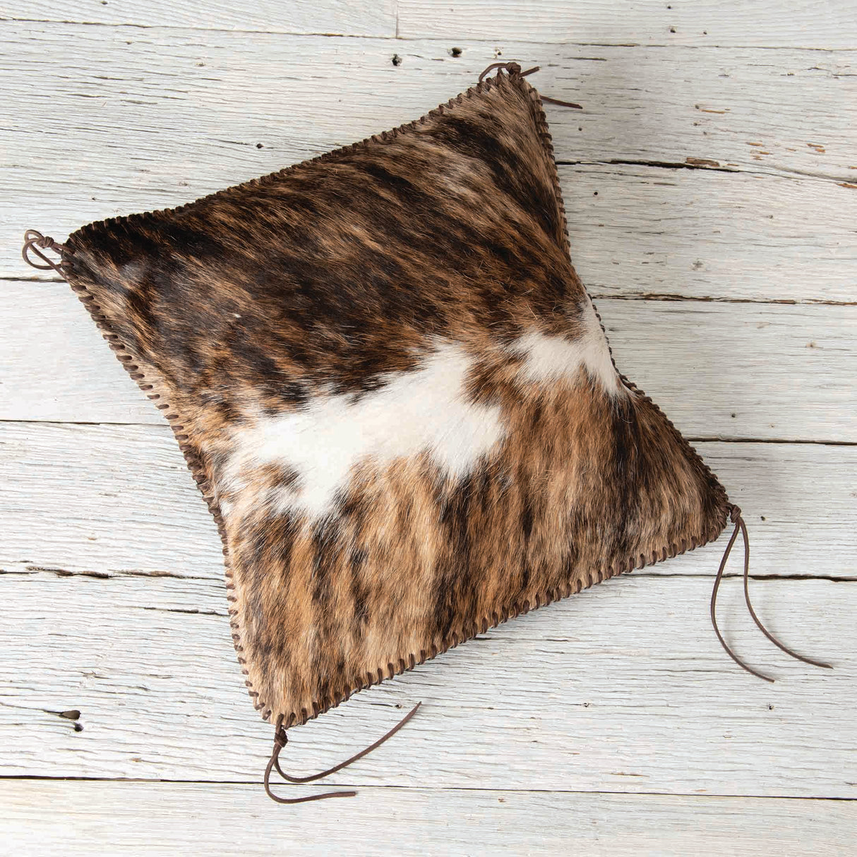 A square decorative cushion with a textured, spotted brown and white fur design, trimmed with brown laces, resting on a rustic wooden surface.