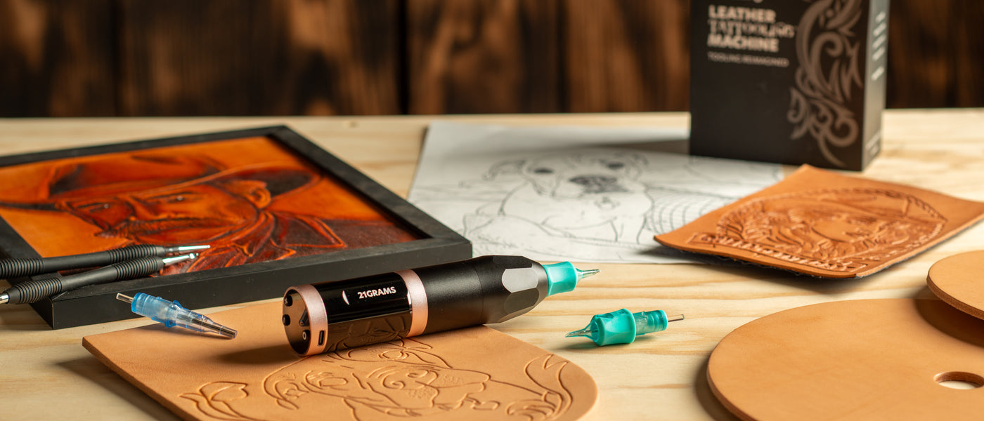 Leather tattooing pen resting on a tooled leather panel on a wooden workbench, with interchangeable needle tips, carved leather artwork, and design sketches in the background.